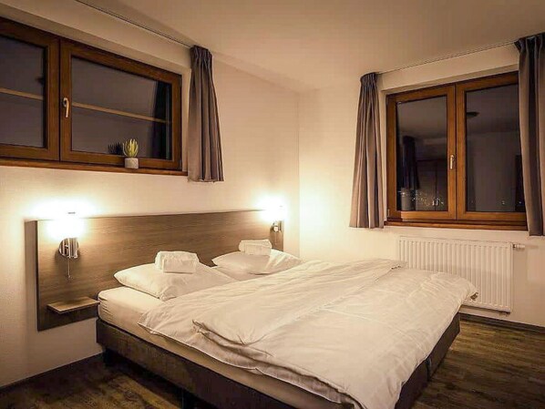 Une chambre d’hôtel avec un lit en bois, deux tables de chevet, une petite table, une fenêtre avec des rideaux et une étagère murale agrémentée d’une plante.