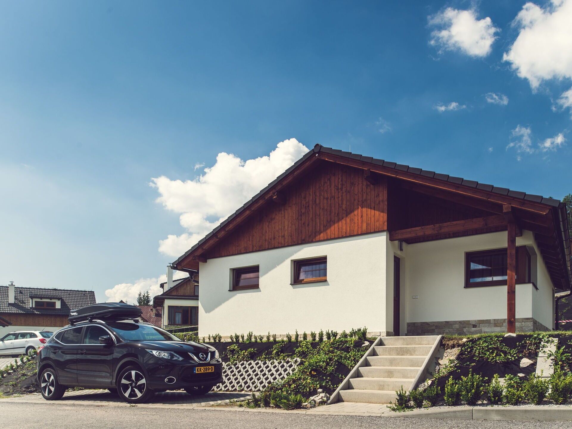 A modern two-story house with a wooden roof and white walls, a car parked in front, and a staircase leading to the entrance.