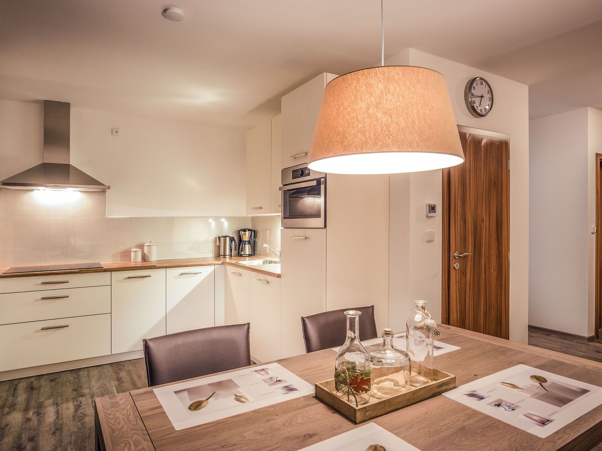 Cocina moderna con armarios blancos, extractor de acero inoxidable y encimera de madera. Hay una mesa de comedor con posavasos y una bandeja con objetos decorativos.