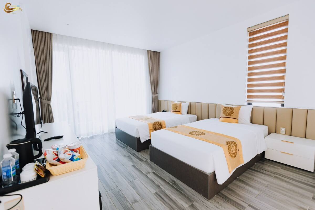 A hotel room with two beds, a TV, a desk with a basket of toiletries, and a window with blinds.