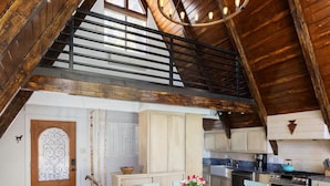 Un loft spacieux avec un plafond haut, des poutres apparentes, un coin repas avec une table blanche et des chaises bleues, et une cuisine avec des meubles blancs et une cuisinière.