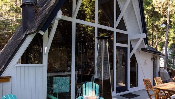 Une cabane dotée d’une grande fenêtre, d’une cheminée et d’un espace extérieur aménagé avec des chaises et un foyer.