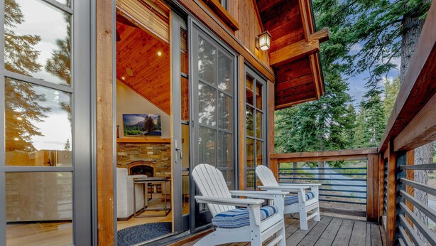 Une terrasse en bois avec des fauteuils Adirondack, une cheminée en pierre et une porte vitrée donnant sur une pièce dont l’un des murs est en pierre et qui est décorée d’un tableau.