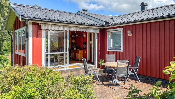 Ein rotes Holzhaus mit Ziegeldach, eine Glastür zu einer Terrasse mit Tisch und Stühlen und ein kleiner Garten.