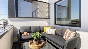 Un balcon moderne avec un canapé d’angle gris, un panier tressé contenant une plante et un chapeau posé sur l’assise.