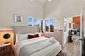 A bedroom with a large bed, a bedside lamp, and a view of the city through windows. - The Conway (Nashville)