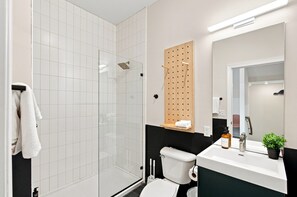 A modern bathroom with a glass shower enclosure, a toilet, a sink with a mirror, and a small shelf with a towel. - The Conway (Nashville)