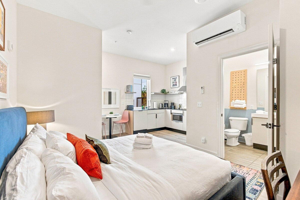 A modern hotel room with a bed, a small kitchenette, and a bathroom.