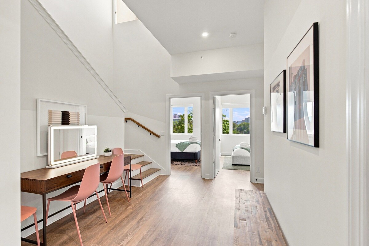A modern interior with a staircase leading to a bedroom, a dining area with pink chairs, and framed artwork on the walls.