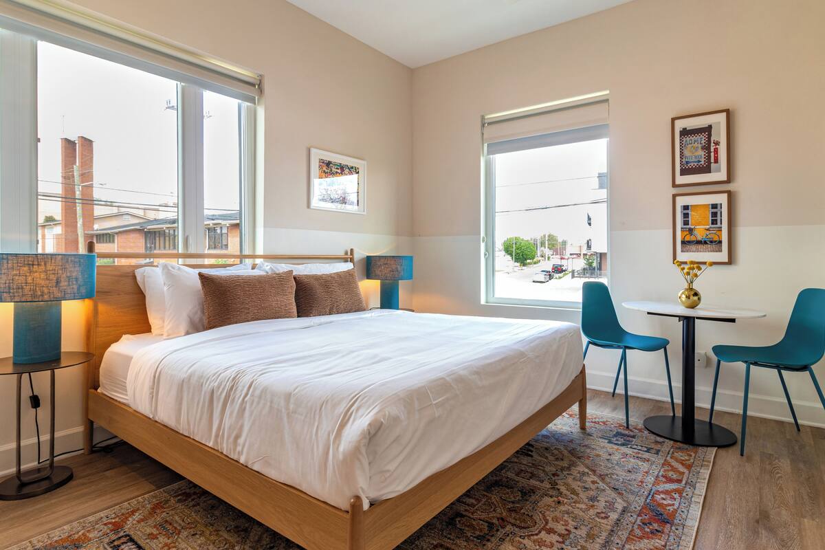 A modern hotel room with a large bed, two bedside tables, a small round table with chairs, and large windows offering a city view.