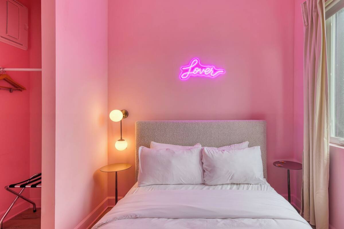 A pink-themed hotel room with a bed, a neon 'Love' sign, a round table, and a window with a view.