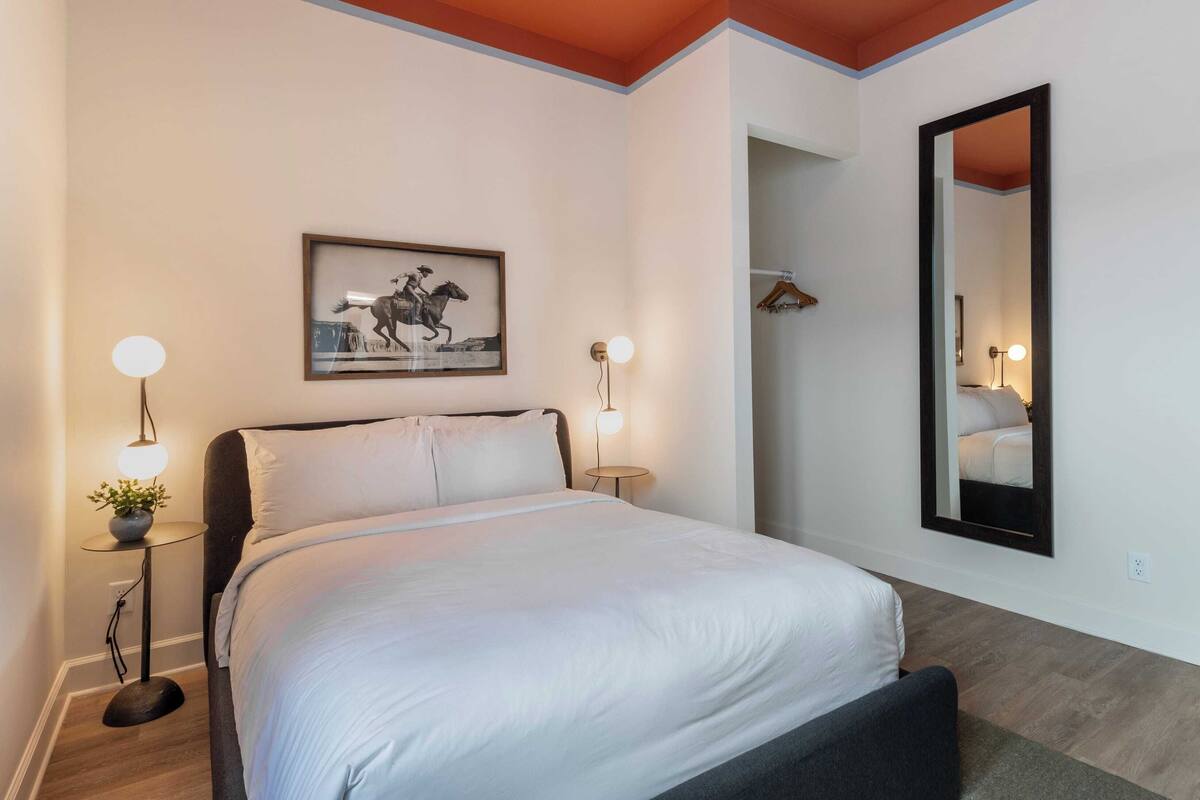 A neatly made bed in a hotel room with a framed picture above it, two bedside lamps, and a large mirror on the wall.