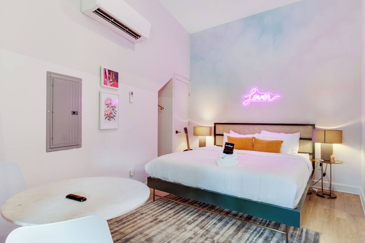 A modern bedroom with a bed, bedside tables, a small round table, and a neon sign on the wall.