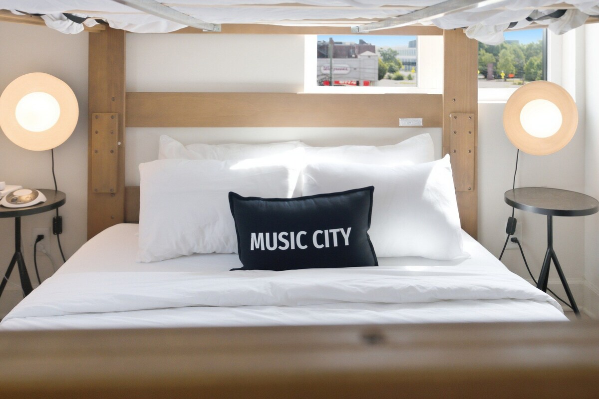 A hotel room with a bed, two bedside tables, two wall-mounted lamps, and a window with a view of the outside.