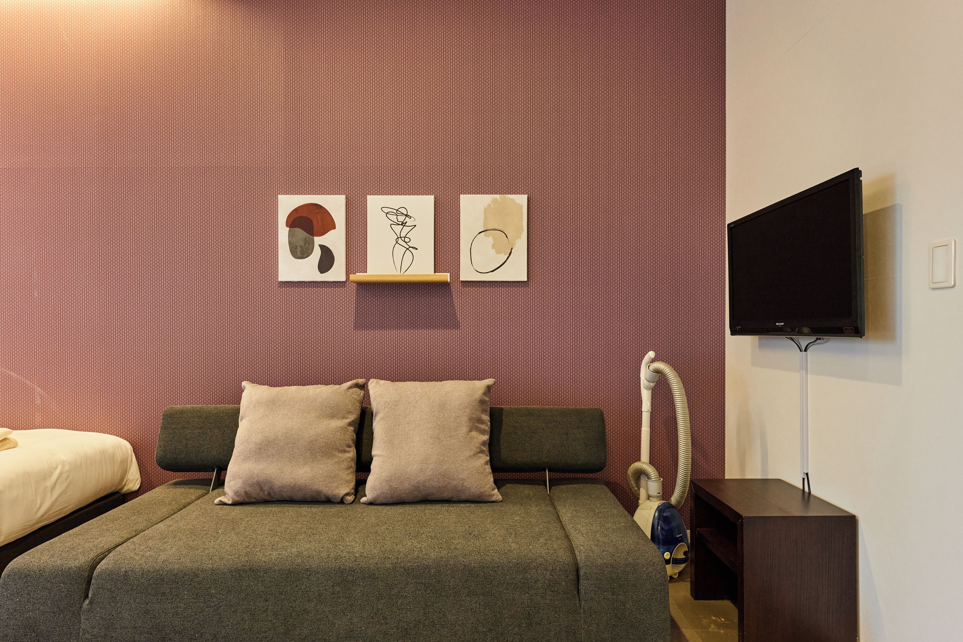 A modern hotel room with a grey sofa bed, a small bedside table, a flat-screen TV mounted on the wall, and abstract wall art.