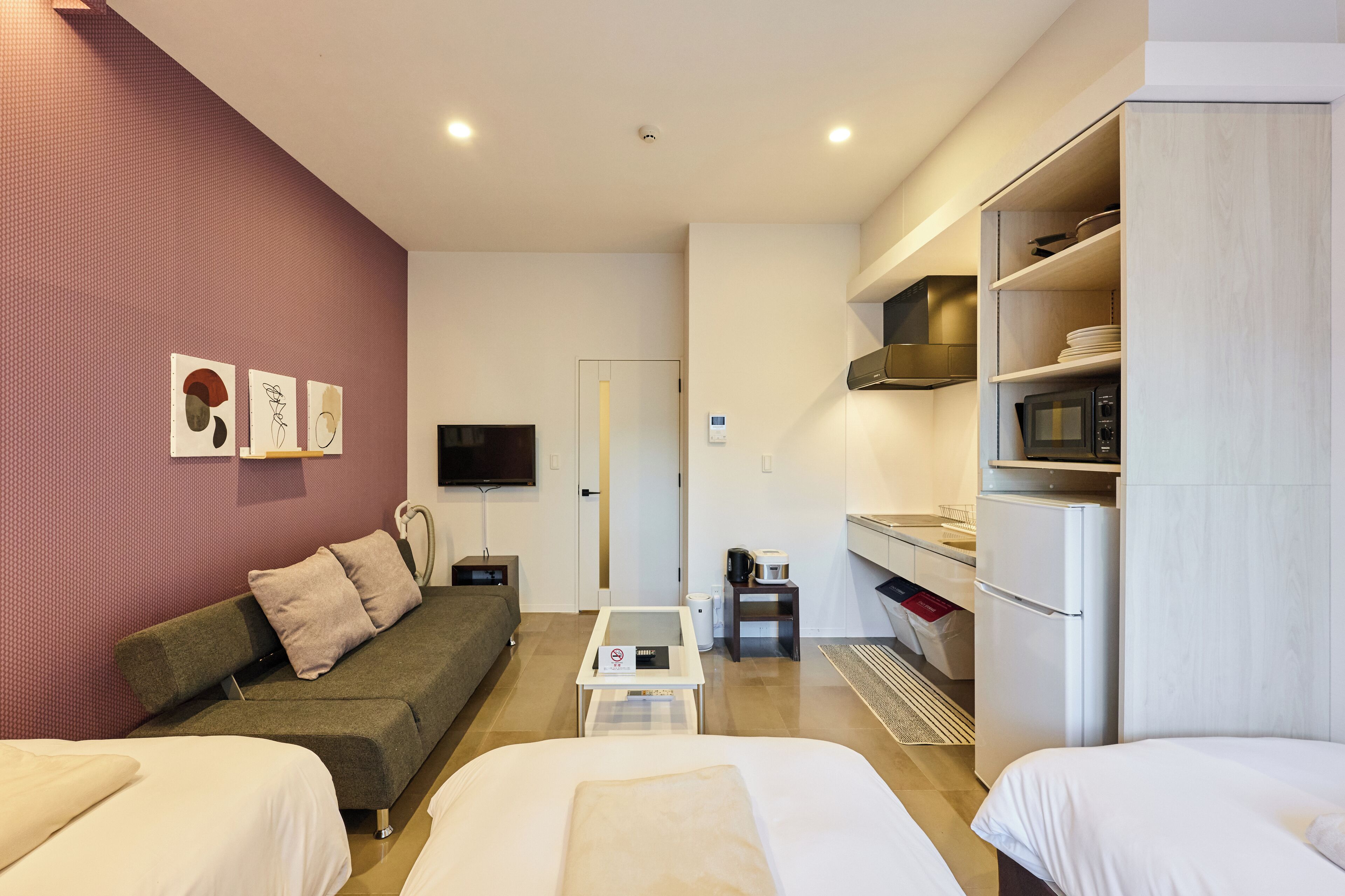 A modern hotel room with a sofa, two beds, a dining table, a kitchen area with a microwave, and a TV.