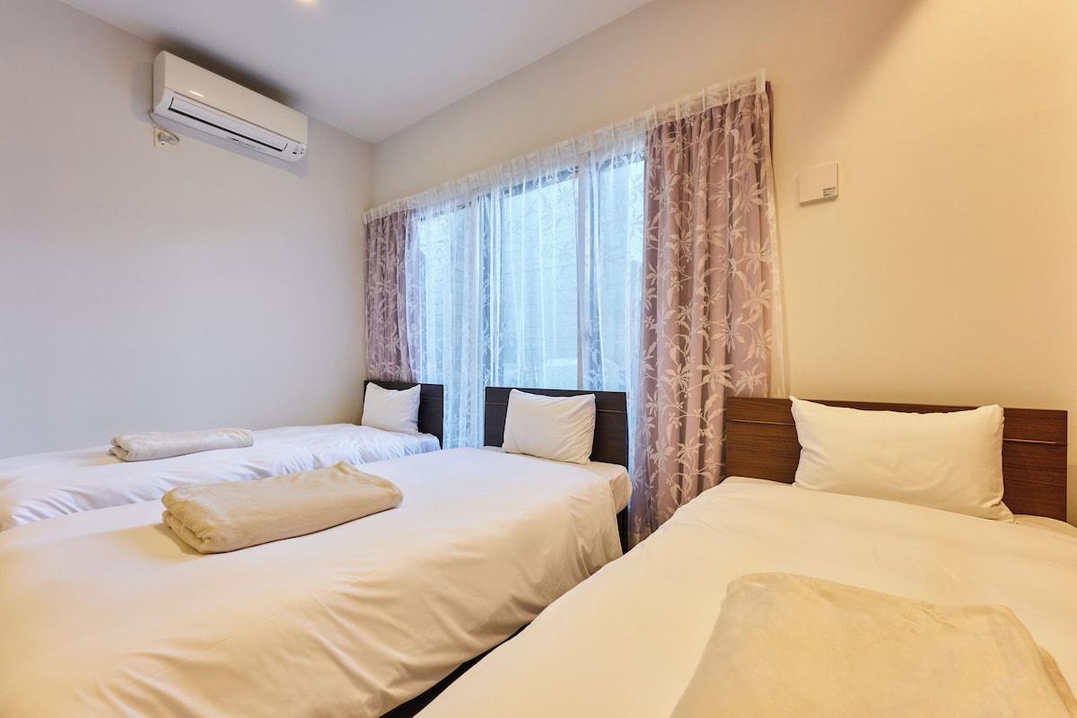 A hotel room with two beds, a window with sheer curtains, an air conditioning unit, and a thermostat on the wall.