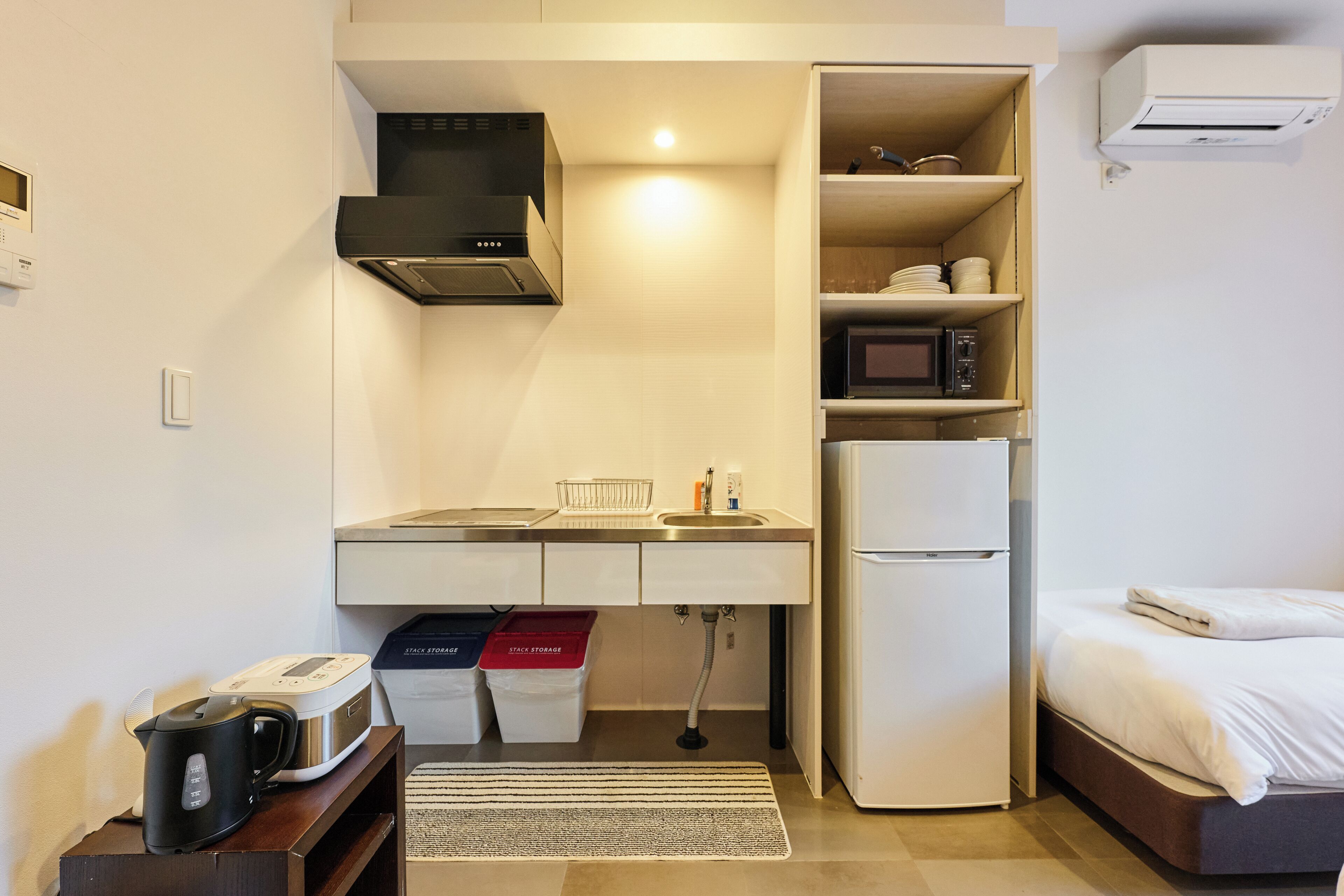 A compact kitchenette with a microwave, stove, sink, and storage cabinets. A refrigerator is adjacent to the kitchenette, and a bed is visible in the background.