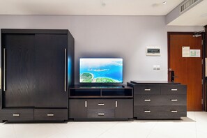 A modern living room with a large flat-screen TV on a wooden entertainment unit, a dark wardrobe, and a wooden door with a notice on it. - The Lancaster Luxury Citypoint ANGIA By An Gia Hospitality (Ho Chi Minh City)