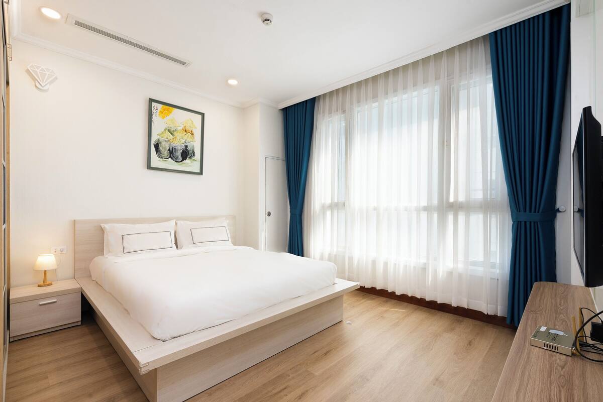 Ein modernes Hotelzimmer mit einem großen Bett, einem Nachttisch mit Lampe, einem Flachbildfernseher und einem großen Fenster mit durchscheinenden Vorhängen.