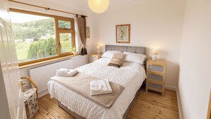 Un dormitorio con una cama grande, suelo de madera, una ventana con vistas a zonas verdes, una mesita de noche con una lámpara y un radiador.