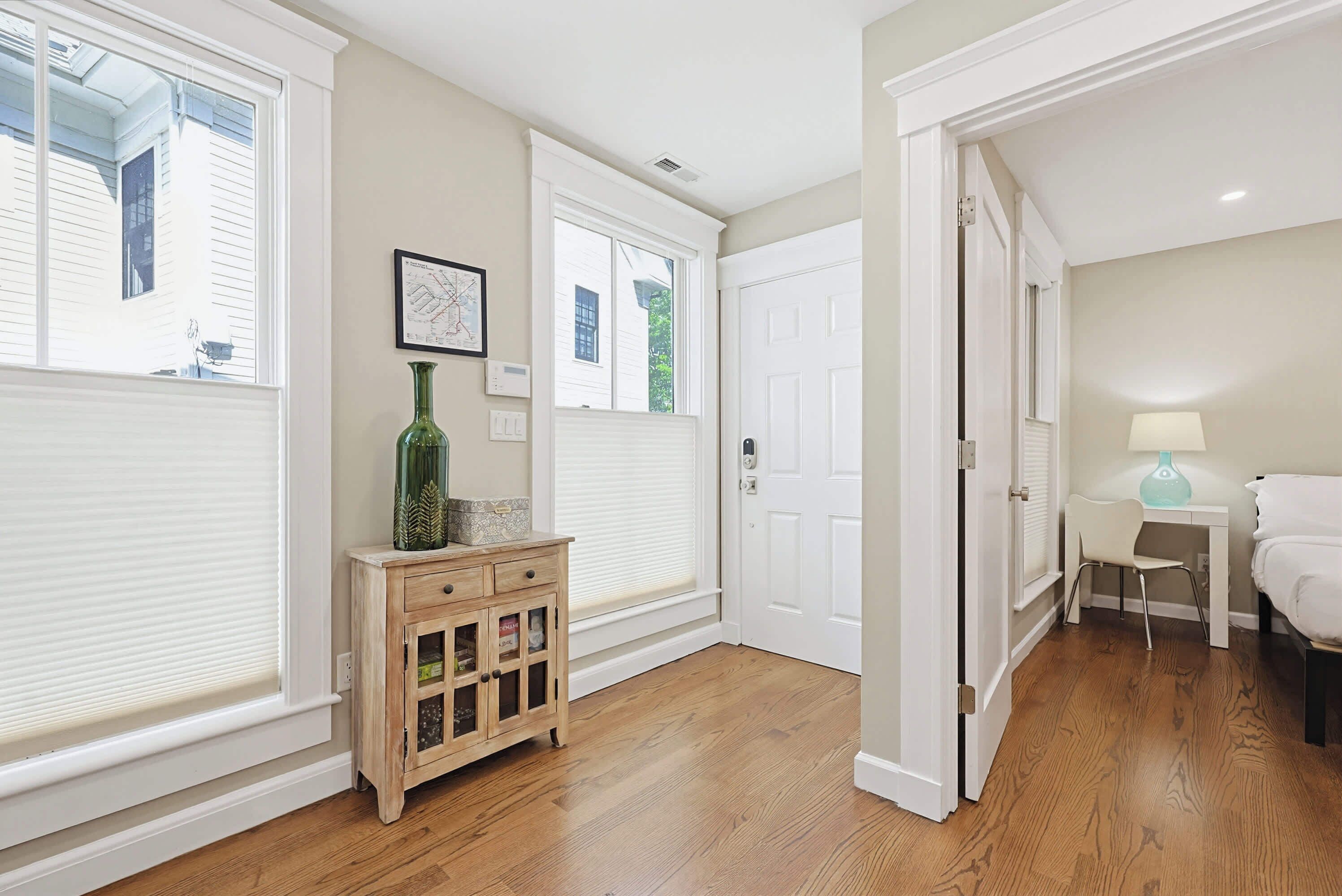 A hallway with wooden flooring, a small cabinet, a door, and a window with blinds.