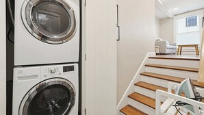 A laundry area with stacked washing machines, a staircase leading to a living area with a couch, and a window. - Inviting 3BR In Cambridge, Walk To T & Central Sq (Cambridge)