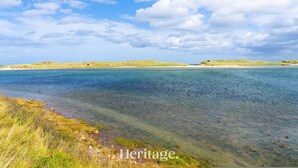 Un paisaje costero con agua cristalina, dunas cubiertas de hierba y una línea de costa distante.