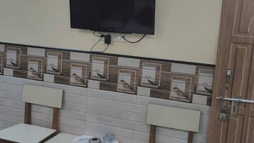 Une petite pièce bien éclairée, avec un lit, un téléviseur fixé au mur, un mur recouvert de carreaux décorés de motifs d'oiseaux, deux chaises en bois et une petite table sur laquelle se trouvent une télécommande et des verres.