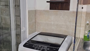 Een compacte, witte wasmachine in een kleine keuken met een betegelde spatwand, een houten plank met een potplant en een raam met uitzicht.