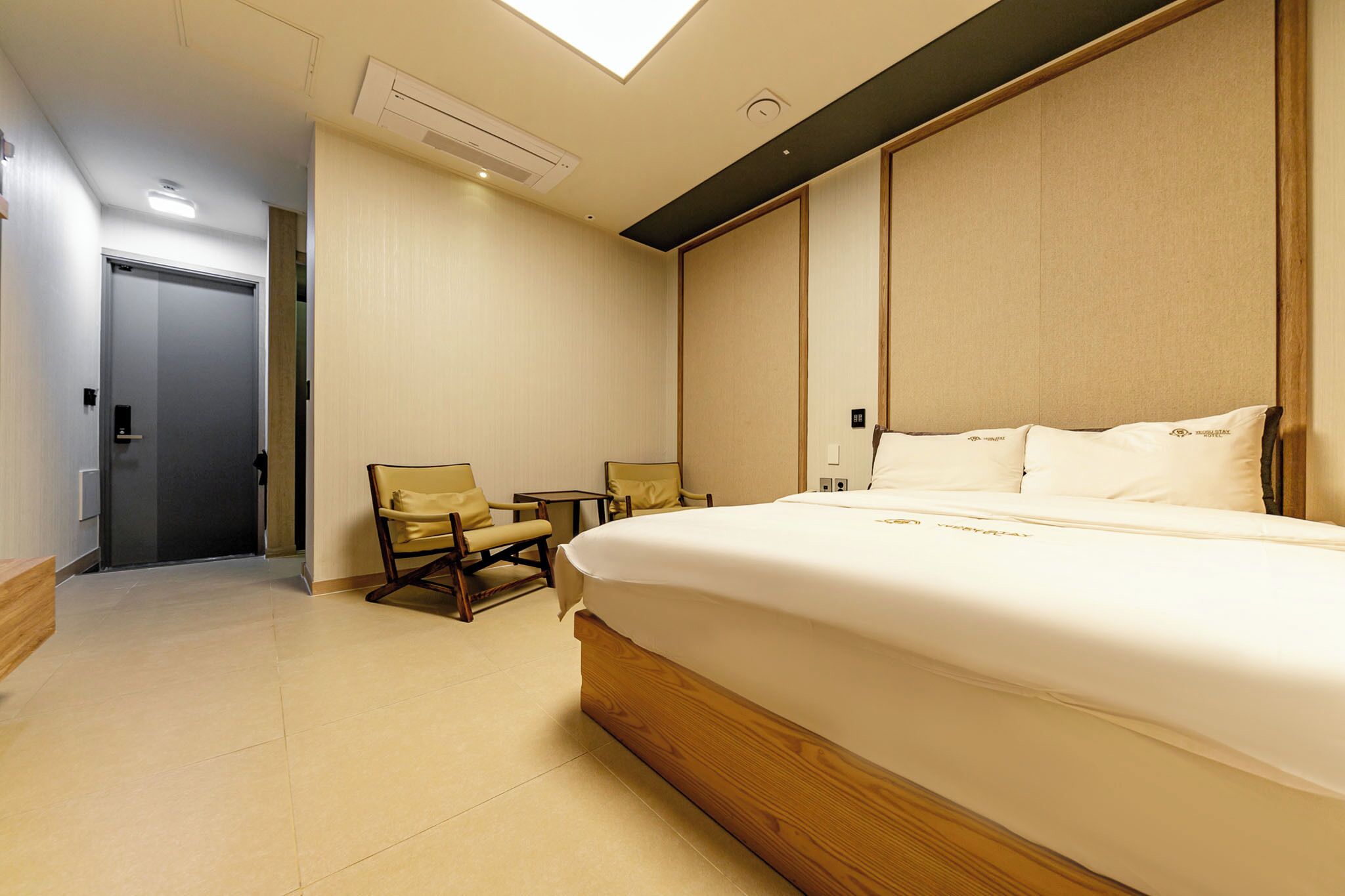 A hotel room with a large bed, a chair, a small table, and a door leading to another room.