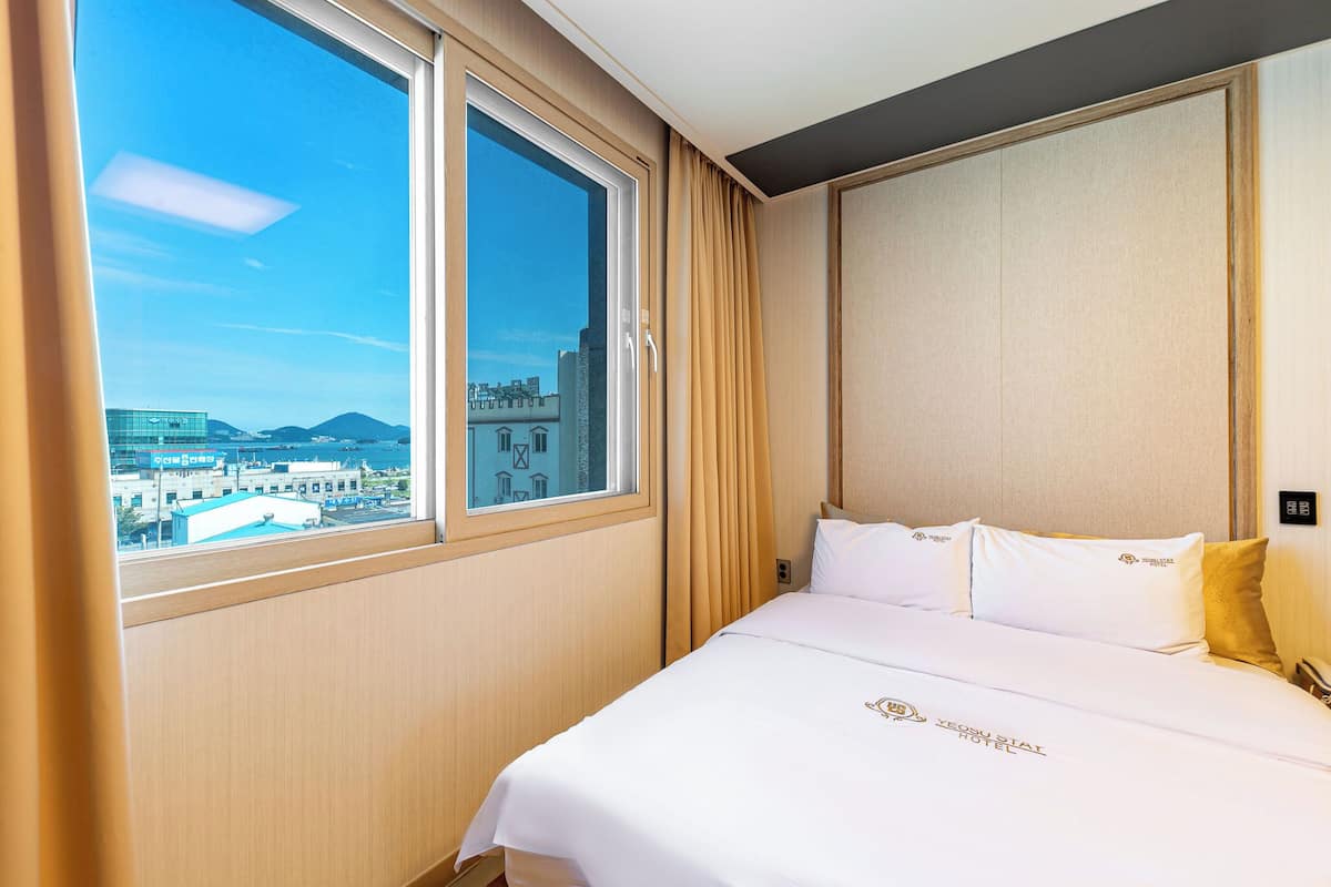 Habitación de hotel con un ventanal grande, cama con ropa de cama blanca y vistas a edificios y montañas.