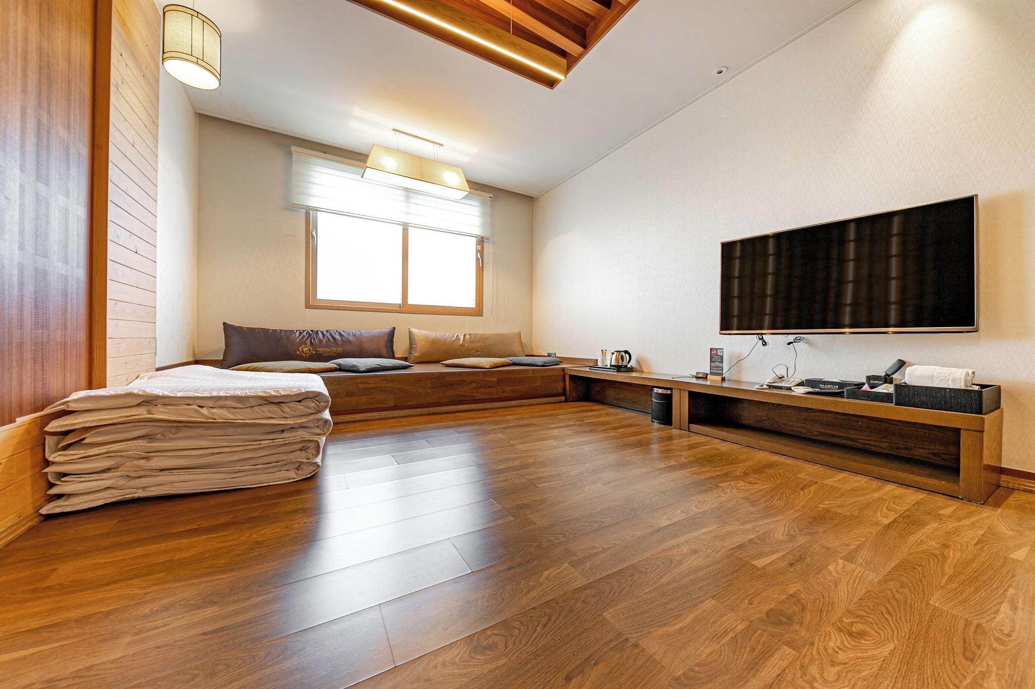 A modern hotel room with a wooden floor, a bed with a beige blanket, a bench with pillows, a flat-screen TV mounted on the wall, and a window with blinds.