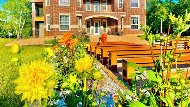 A two-story brick building with a central entrance, surrounded by a garden with benches and flowering plants.