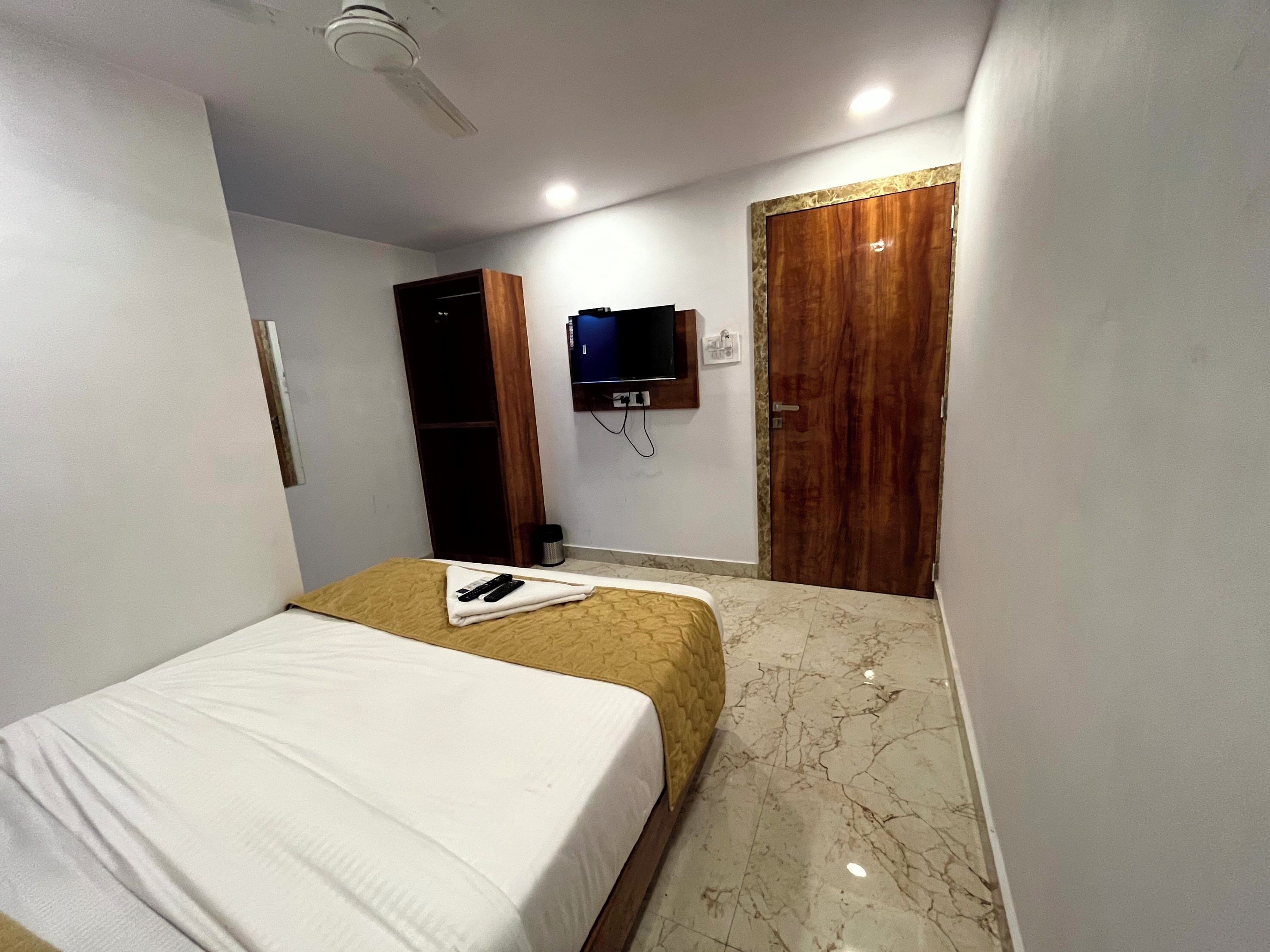 A hotel room with a bed, a television mounted on the wall, a wooden wardrobe, and a ceiling fan.