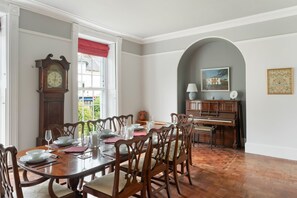 A dining room with a wooden table set for six, a grandfather clock, and a piano in the background.