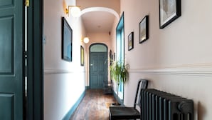 A hallway with a wooden floor, a radiator, a chair, and framed pictures on the walls. - The Emporium - Stylish 2BD in Winchcombe (Cheltenham)