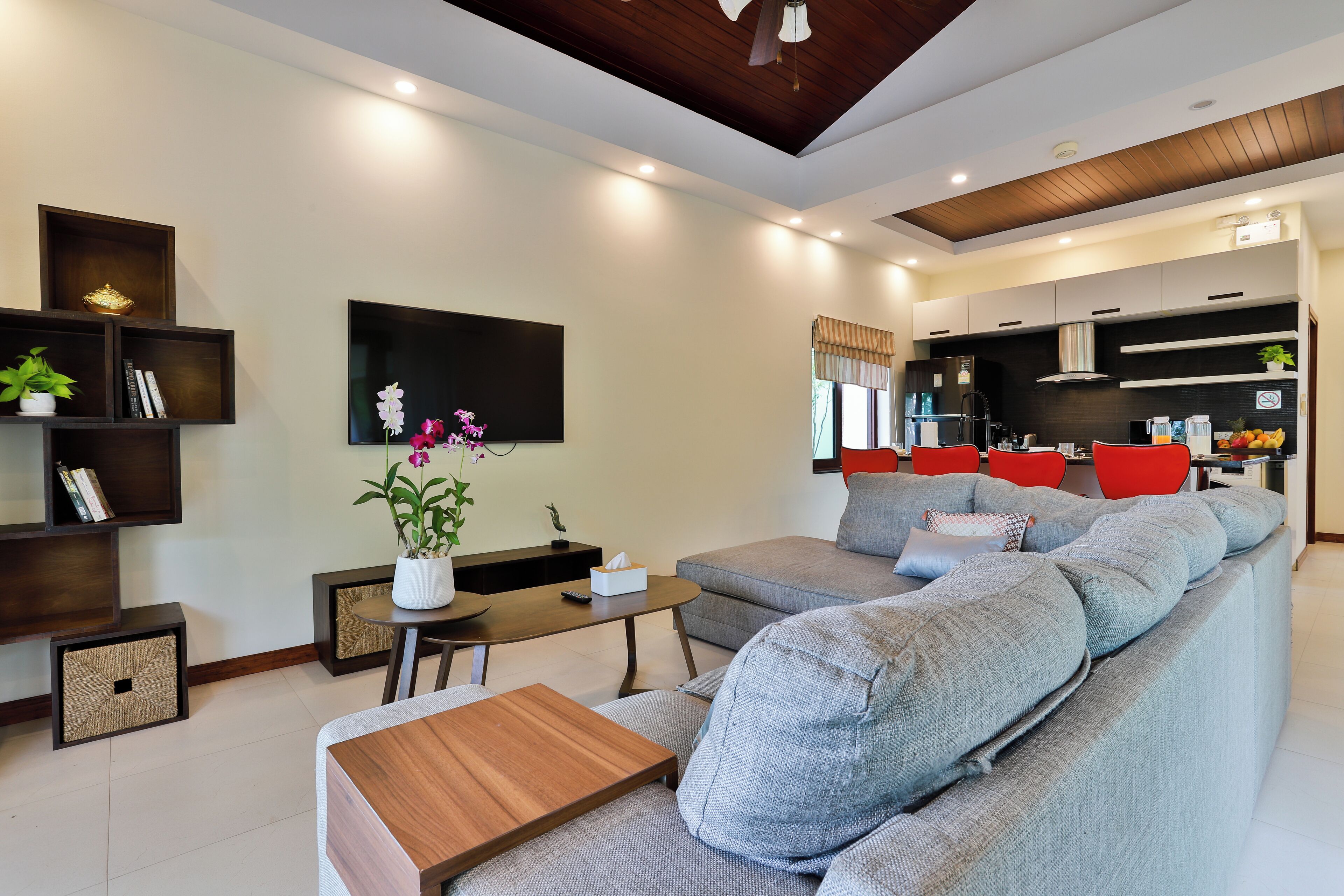 A modern living room with a grey sectional sofa, a wooden coffee table, and a TV mounted on the wall. There is a kitchen area with red chairs and a dining table.