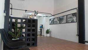 Une pièce spacieuse avec un plancher en bois, une chaise noire, un bureau noir et une plante en pot. Trois œuvres d’art encadrées ornent le mur, et un luminaire est fixé au plafond.
