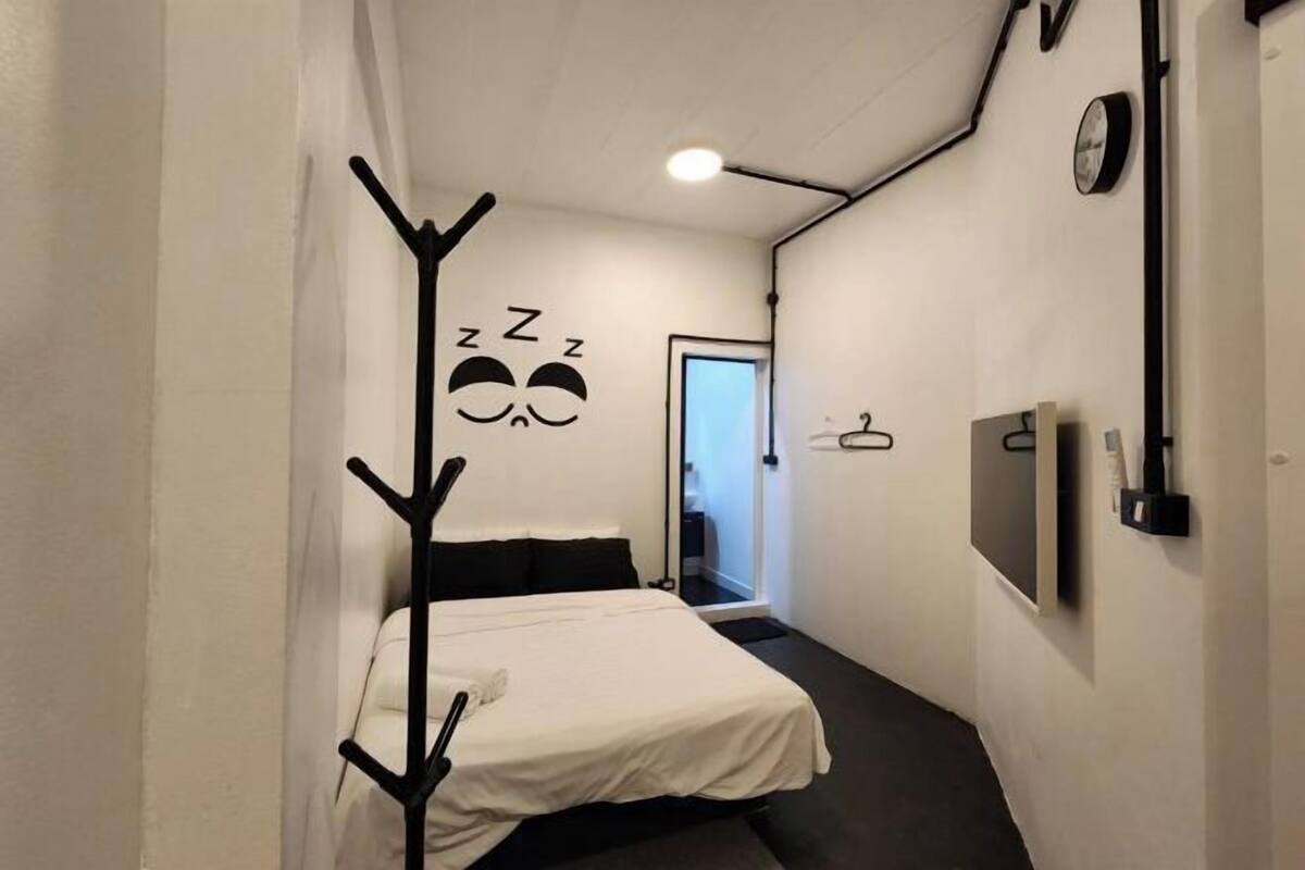 A small, narrow hotel room with a bed, a coat rack, a wall clock, and a television.