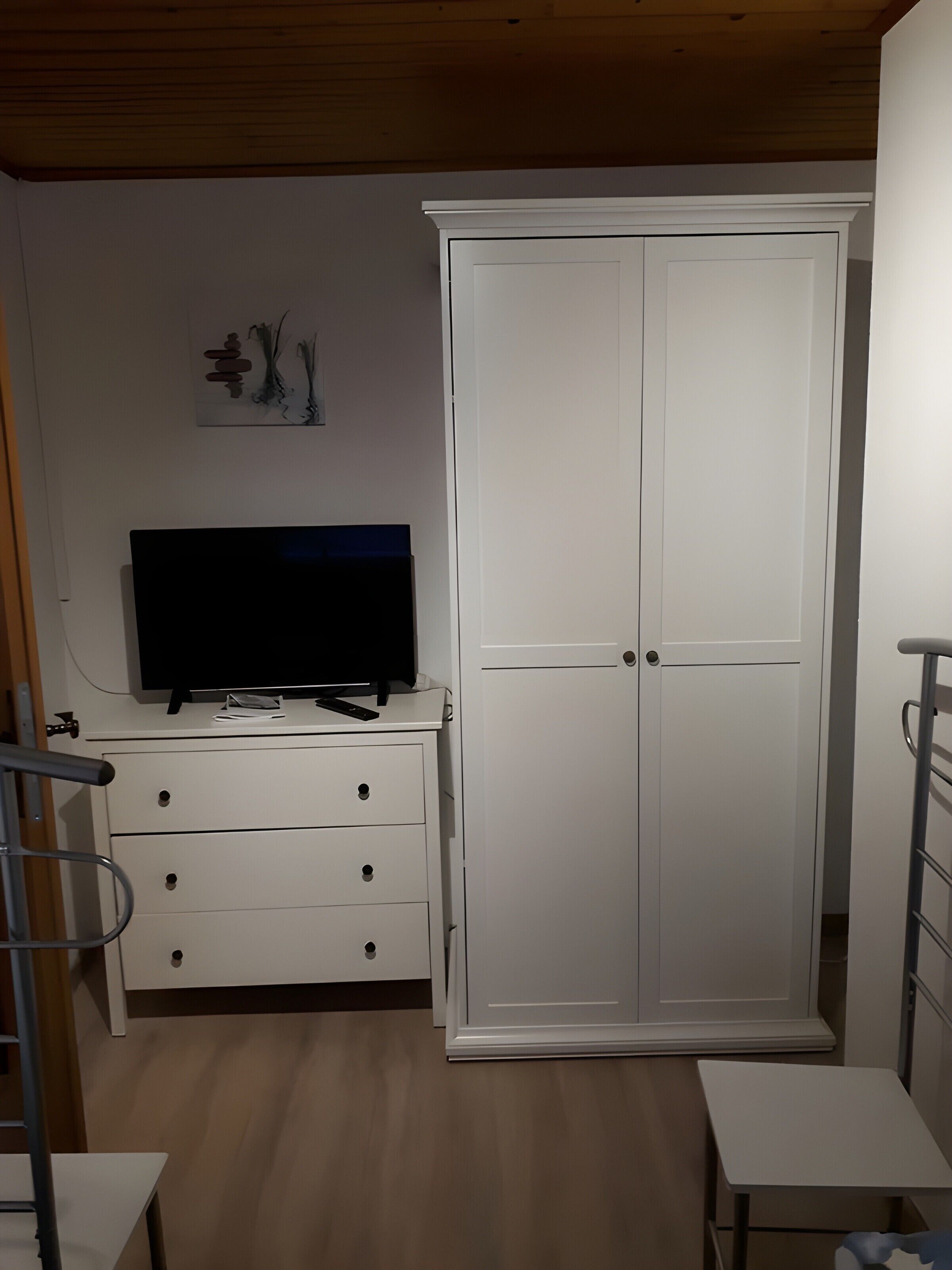 A bedroom with a white wardrobe, a TV on a dresser, a small table, and a metal chair.
