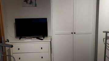 A bedroom with a white wardrobe, a TV on a dresser, a small table, and a metal chair.