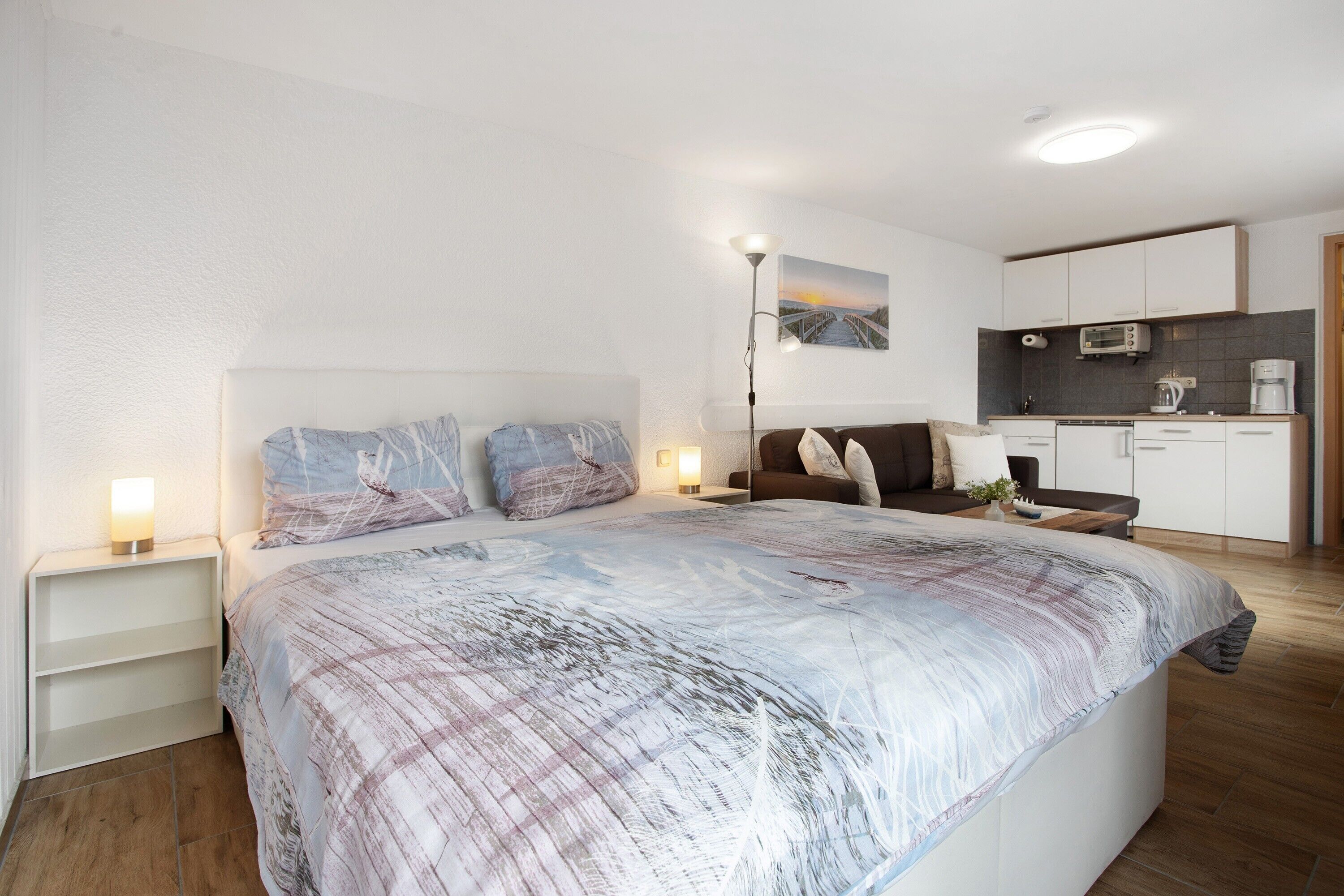 Ein modernes Schlafzimmer mit einem großen Bett, Nachttischlampen, einem Nachttisch, einer kleinen Sitzecke mit einer Couch und einer Küchenzeile im Hintergrund.