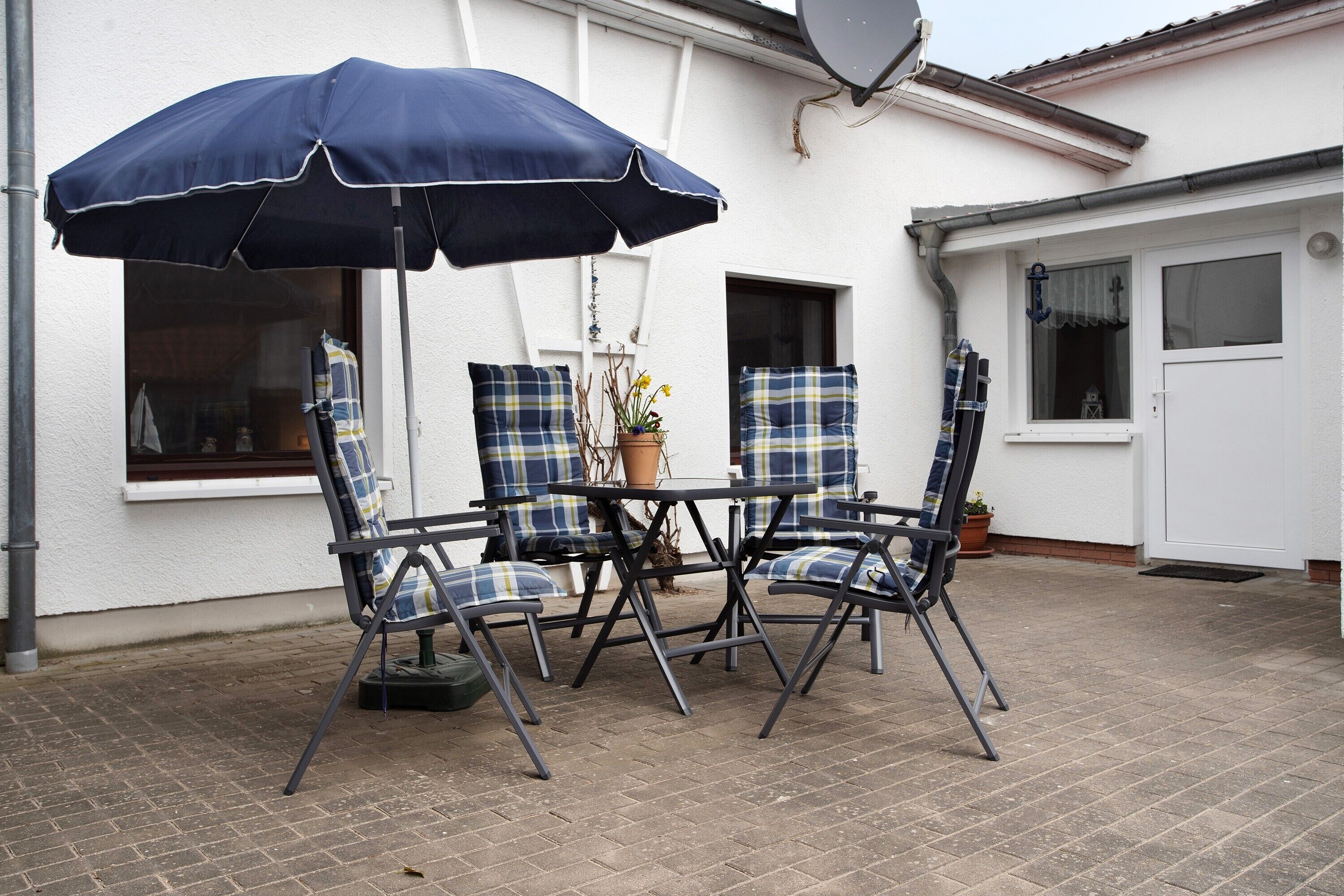 An outdoor patio with a table and four chairs, a blue umbrella, and a potted plant.