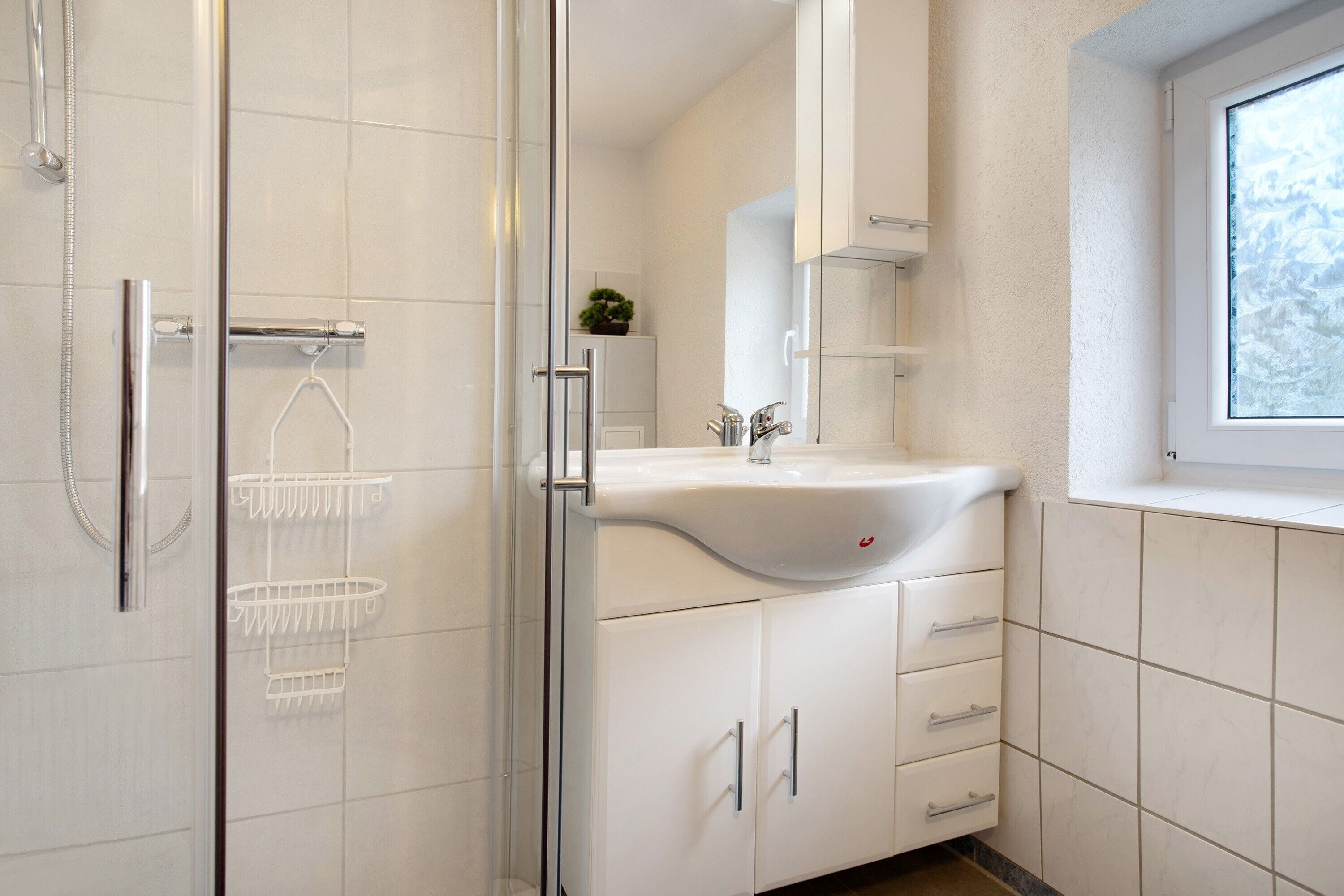 Een moderne badkamer met een glazen douchecabine, witte kastjes en een raam voor natuurlijk licht.