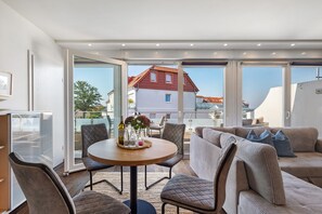 A modern living room with a round table, cushioned seating, and a balcony with a view of a red-roofed building.
