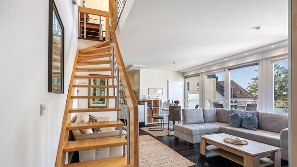 Un interior modern amb una escala de fusta, una zona d'estar amb sofà i taula de centre, i una cuina al fons.