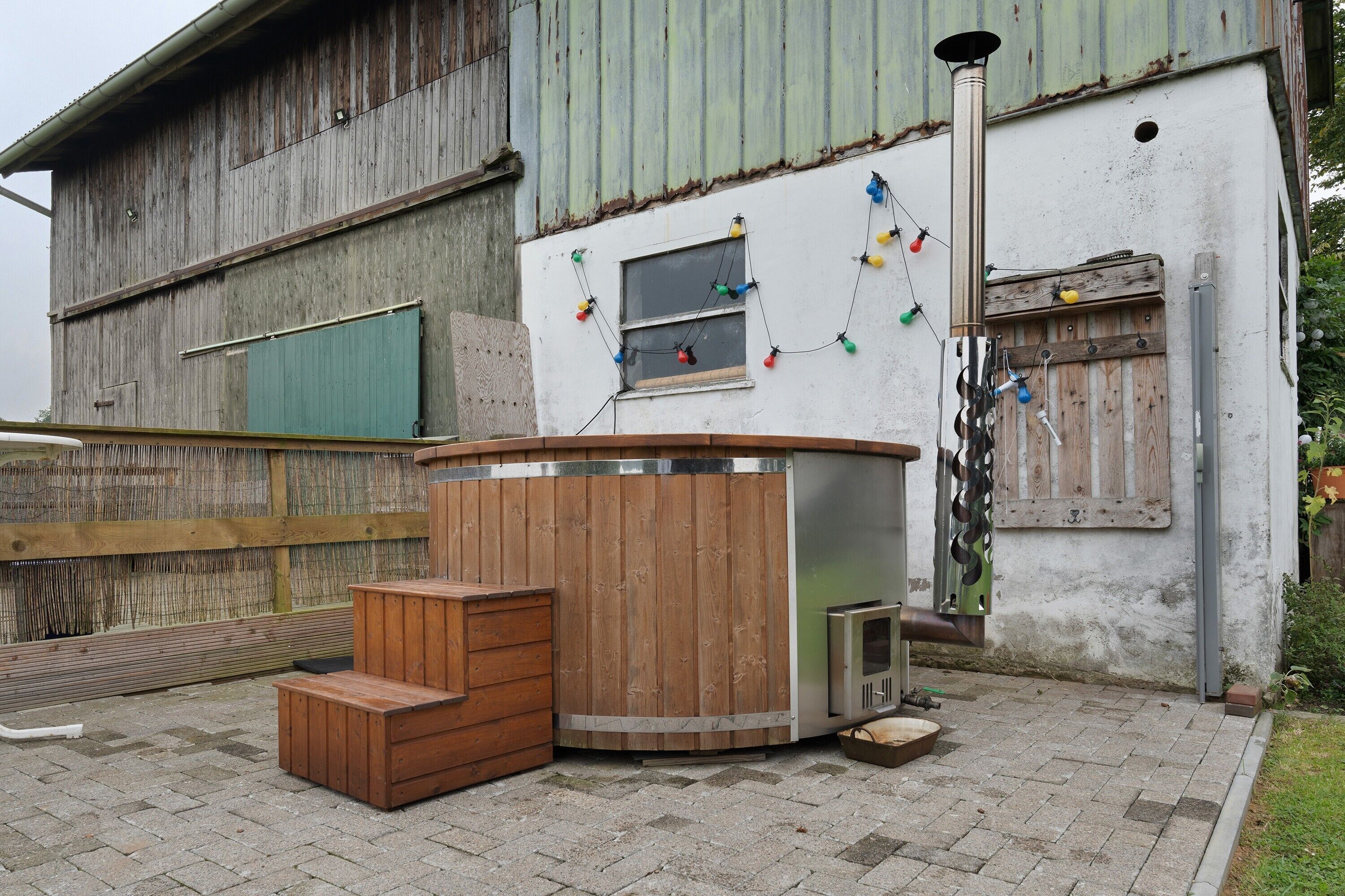 Un'area esterna dall'aspetto rustico con una panchina in legno e un tavolo in metallo, una piccola finestra con luci colorate e un recinto di legno.