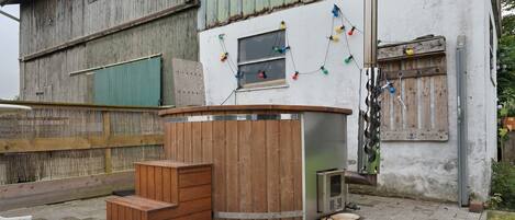 A rustic outdoor seating area with a wooden bench and a metal table, a small window with colorful lights, and a wooden fence.