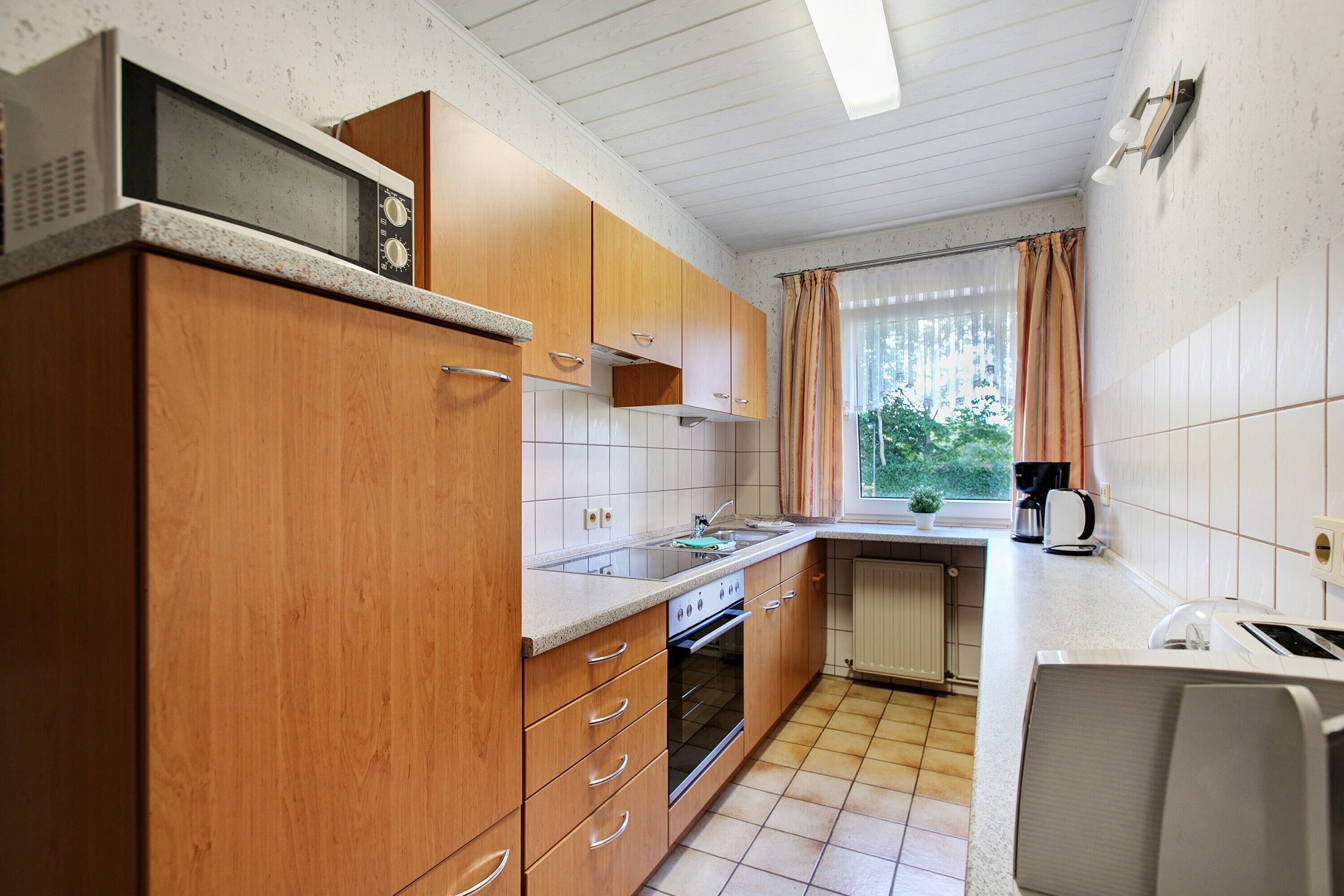 A compact kitchen with wooden cabinets, a built-in microwave, a stove, and a window with curtains.